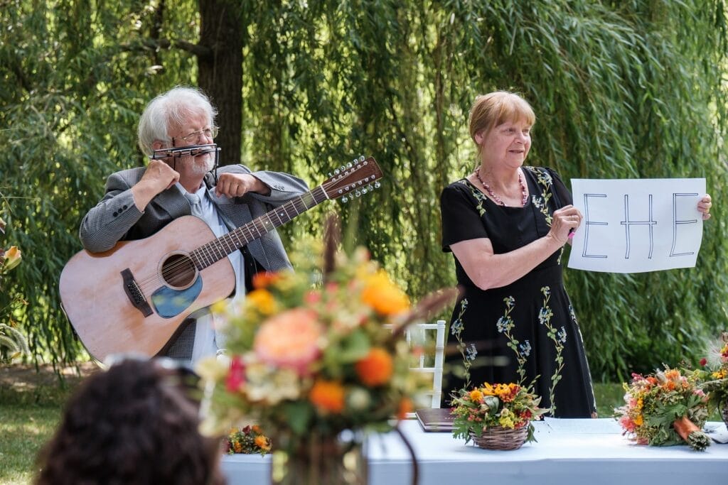 Hochzeitskrone Ein älterer Mann spielt Gitarre und Mundharmonika, während eine ältere Frau ein Schild mit der Aufschrift „EHE“ hochhält und so echte Momente festhält. Sie befinden sich im Freien inmitten wunderschöner Blumendekorationen, die an authentische Hochzeitsfotos eines Hochzeitsfotografen in Wien und Niederösterreich erinnern.