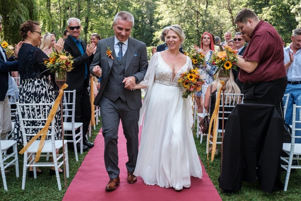 Hochzeitskrone Ein Paar in Hochzeitskleidung geht Hand in Hand einen Gang im Freien entlang, der von Menschen gesäumt ist, die klatschen und authentische Hochzeitsfotos machen. Die Braut hält einen Strauß Sonnenblumen.