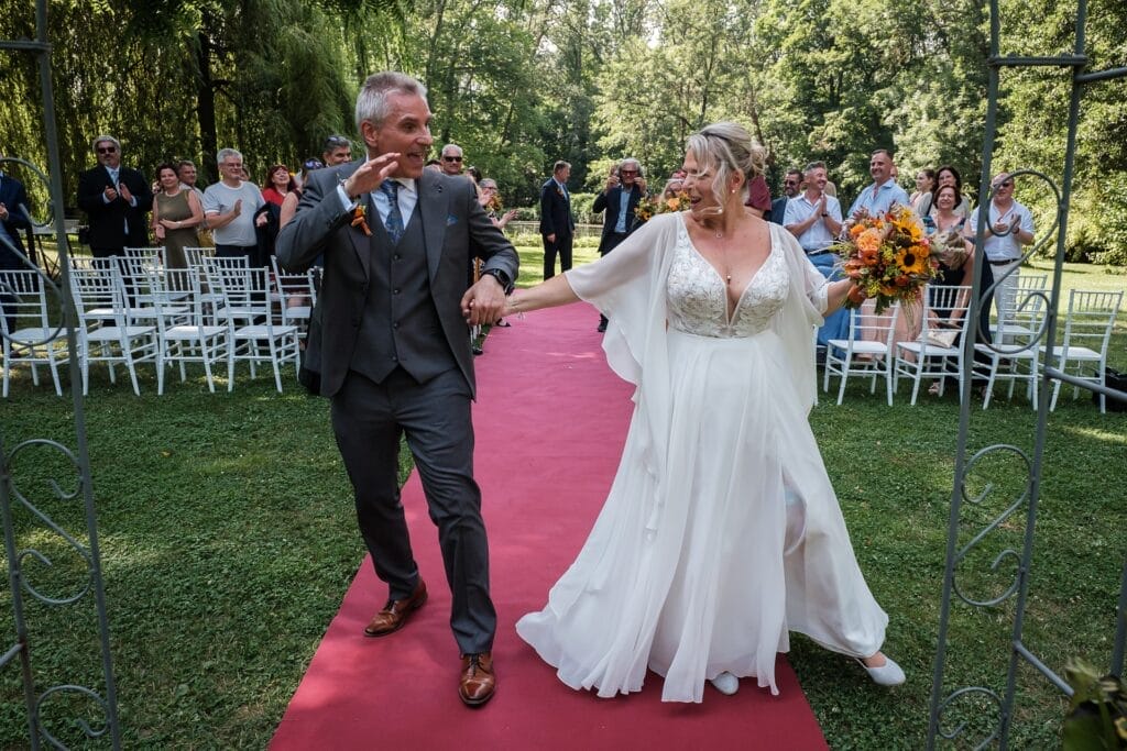 Hochzeitskrone Ein frisch vermähltes Paar schreitet freudig über einen roten Teppich nach draußen, umgeben von klatschenden Gästen. Die Braut hält einen Blumenstrauß in der Hand, beide lächeln – echte Momente, die jeder Hochzeitsfotograf in Wien und Niederösterreich in Erinnerung behalten würde.