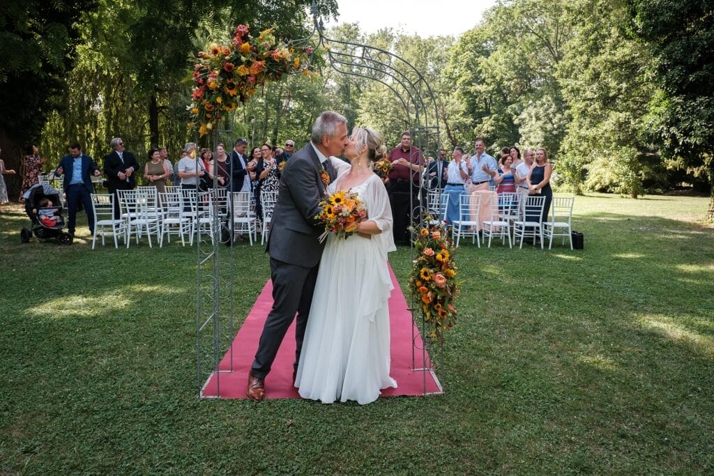 Hochzeitskrone Unter einem Blumenbogen auf einem roten Teppich küssen sich Braut und Bräutigam und halten so *echte Momente* fest. Bei dieser bezaubernden Hochzeit im Freien applaudieren die Gäste. Ein Tag voller *authentischer Hochzeitsfotos*, perfekt für jeden Hochzeitsfotografen in Wien und Niederösterreich.