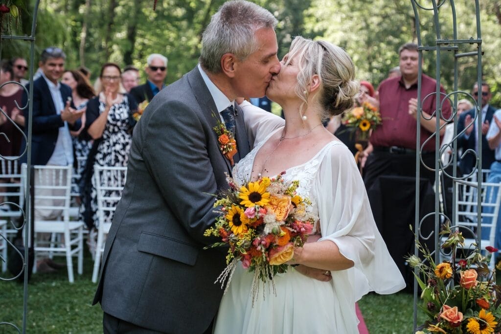 Hochzeitskrone Ein Paar küsst sich bei einer Hochzeitszeremonie im Freien und hält einen Strauß bunter Blumen in der Hand. Gäste im Hintergrund klatschen und verfolgen das Geschehen. Der Hochzeitsfotograf in Wien und Niederösterreich fängt diese echten Momente als authentische Hochzeitsfotos ein, die man für immer in Erinnerung behalten wird.