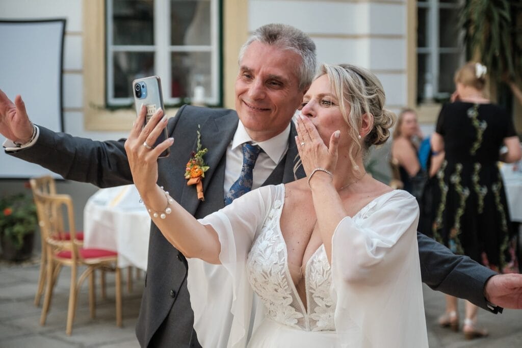 Hochzeitskrone Ein Paar im Hochzeitskleid macht ein Selfie im Freien und fängt echte Momente ein, während die Frau einen Kuss zuwirft.