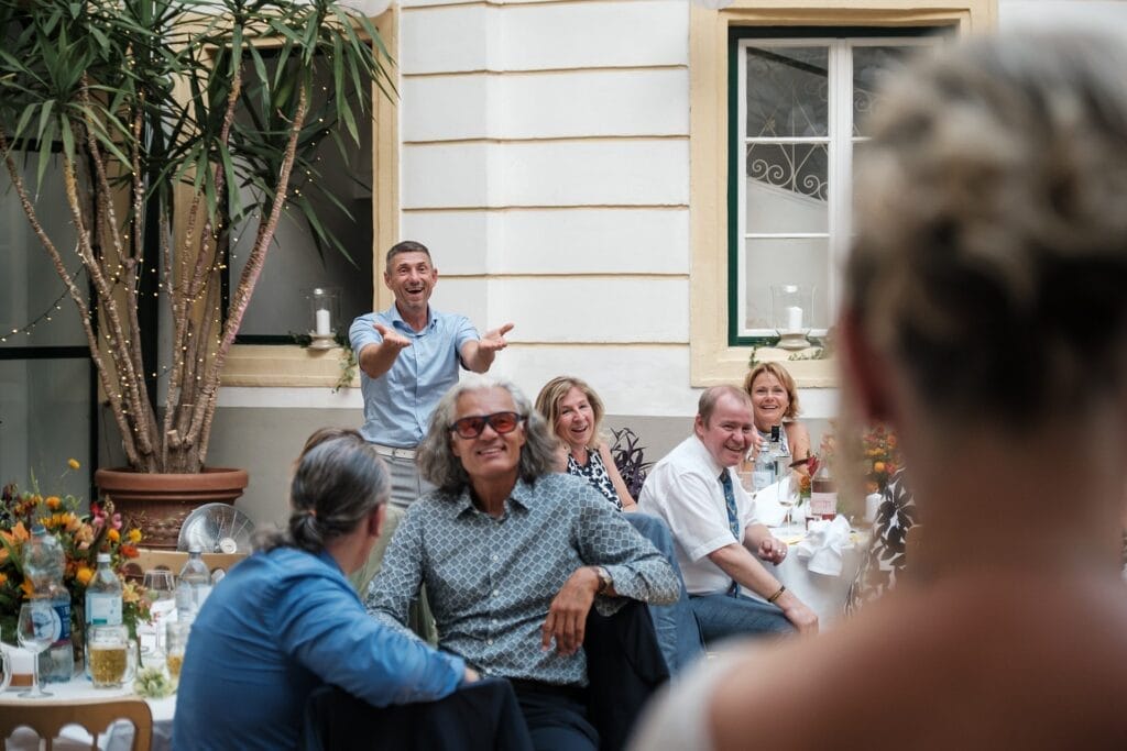 Hochzeitskrone Menschen sitzen an einem Tisch, lächeln und klatschen und machen authentische Hochzeitsfotos bei einer Zusammenkunft im Innenbereich, die mit Pflanzen und dekorativen Lichtern geschmückt ist, und fangen so echte Momente perfekt ein.