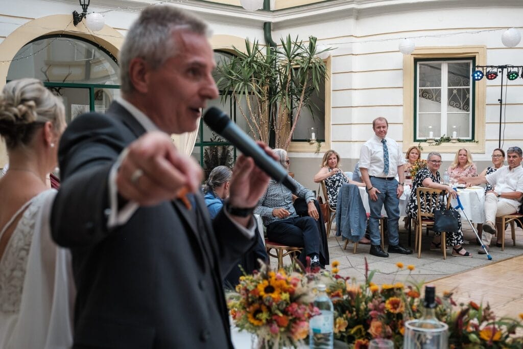 Hochzeitskrone Ein Mann spricht bei einer Versammlung in einem Innenraum in ein Mikrofon und fängt „echte Momente“ ein, während sitzende und stehende Teilnehmer zuschauen. Im Vordergrund ist ein Blumenarrangement zu sehen.