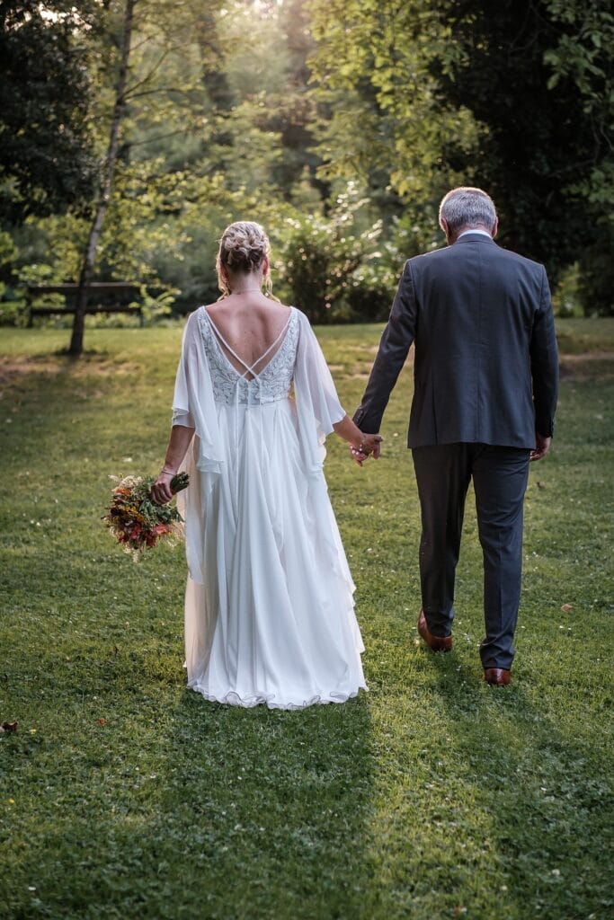 Hochzeitskrone Ein Paar in formeller Kleidung geht Hand in Hand durch einen sonnenbeschienenen Garten, aufgenommen von einem Hochzeitsfotografen in Wien und Niederösterreich. Die Frau hält einen Blumenstrauß und trägt ein weißes Kleid, während der Mann einen grauen Anzug trägt. So entstehen authentische Hochzeitsfotos voller echter Momente.