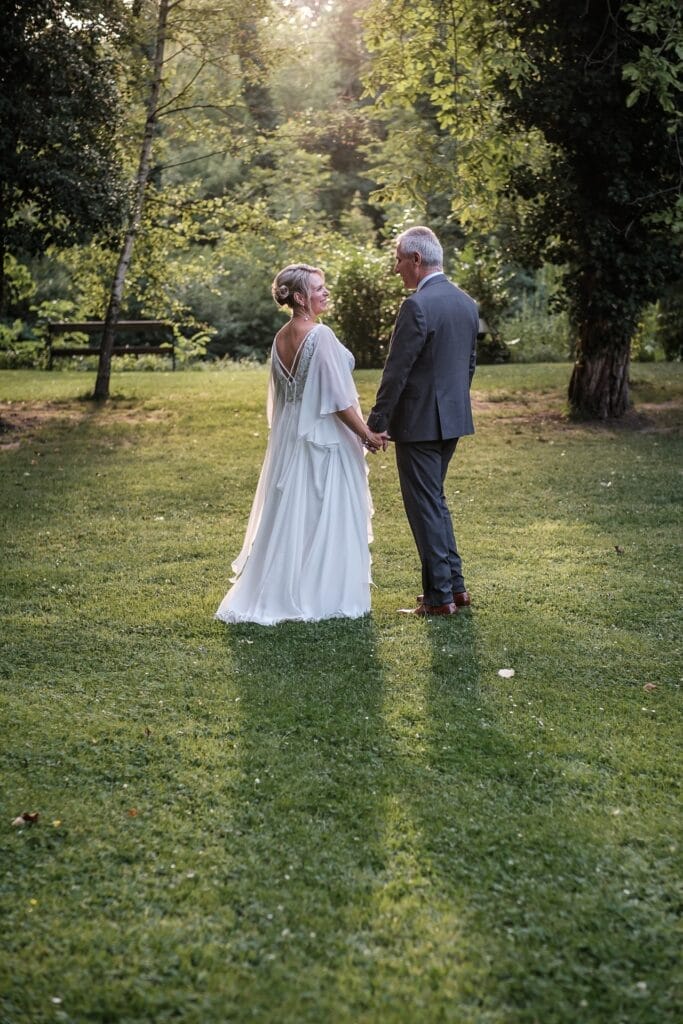 Hochzeitskrone Ein älteres Paar in formeller Kleidung steht Hand in Hand auf einer Wiese, umgeben von Bäumen, durch deren Zweige Sonnenlicht fällt – und fängt echte Momente in authentischen Hochzeitsfotos eines Hochzeitsfotografen aus Wien und Niederösterreich ein.