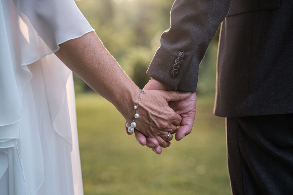 Hochzeitskrone Ein Paar, das sich in formeller Kleidung an den Händen hält, strahlt Eleganz aus. Die Person auf der linken Seite trägt ein weißes Oberteil mit Perlenarmband, während die Person auf der rechten Seite einen dunklen Anzug trägt. Diese Szene fängt echte Momente ein und eignet sich perfekt für authentische Hochzeitsfotos vor einer bezaubernden Kulisse im Freien.