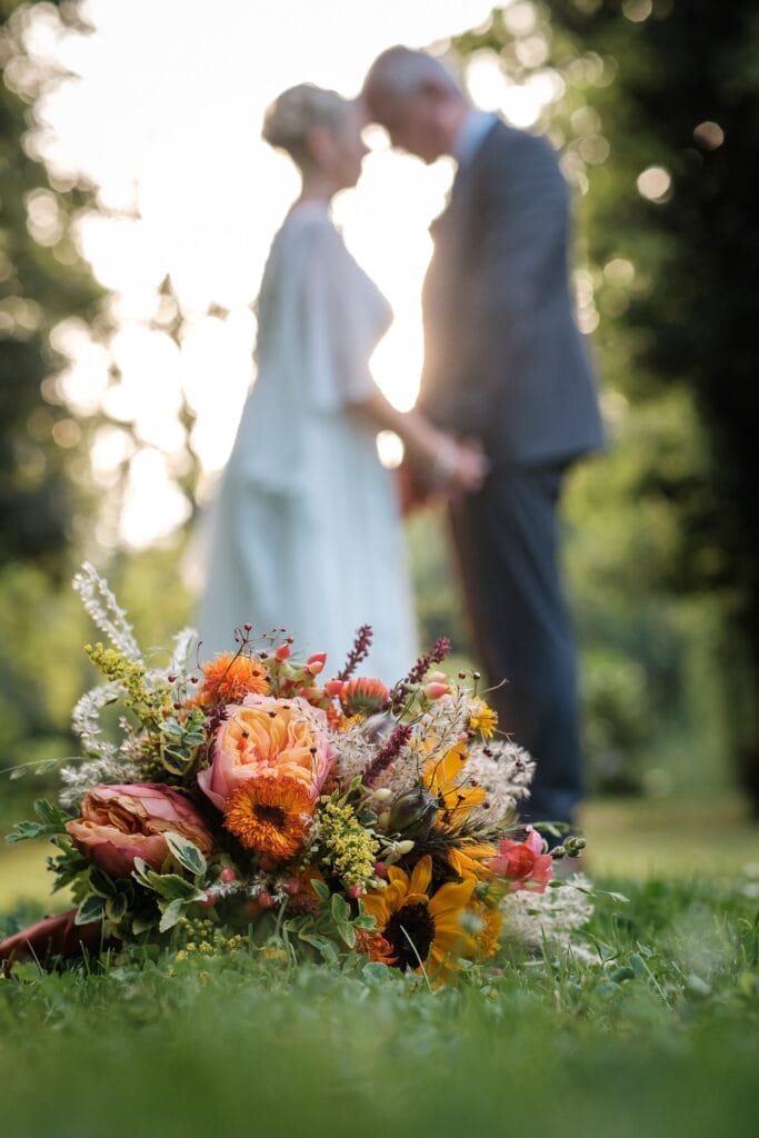 Hochzeitskrone Blumenstrauß auf dem Gras mit einem verschwommenen Paar im Hochzeitskleid, das im Hintergrund seine Stirnen berührt und echte Momente perfekt einfängt.