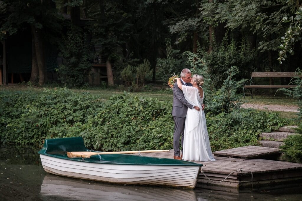 Hochzeitskrone Ein Paar umarmt sich auf einem Holzsteg neben einem kleinen Ruderboot, umgeben von üppigem Grün, und fängt echte Momente der Liebe in einer authentischen Umgebung ein.