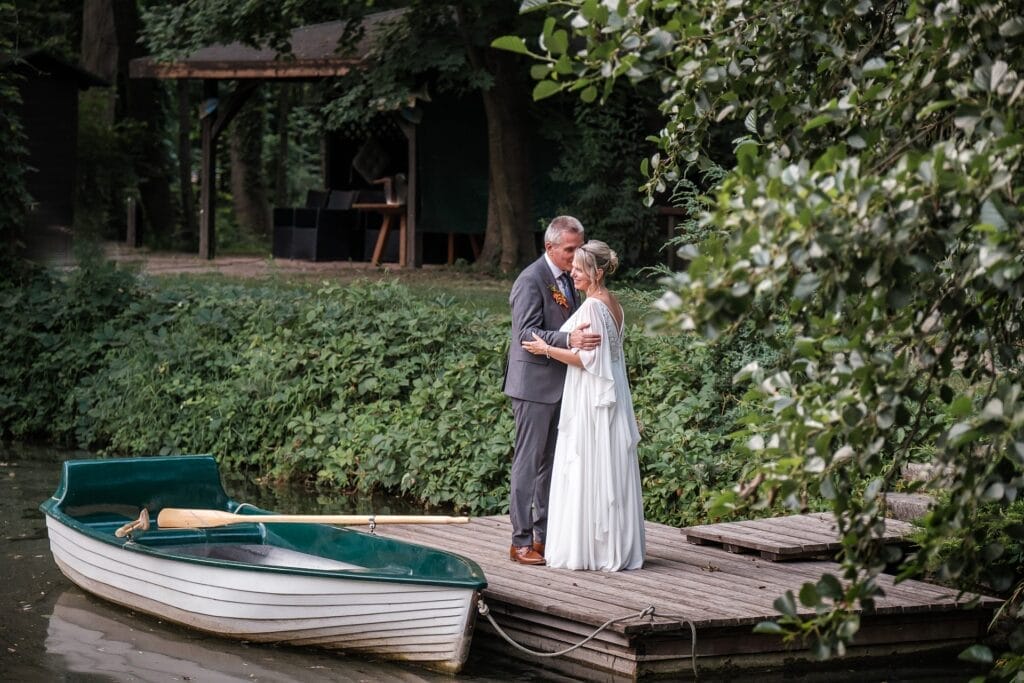 Hochzeitskrone Ein Paar in Abendgarderobe steht auf einem Holzsteg neben einem weißen Ruderboot auf einem ruhigen See, umgeben von Grün, und fängt die Essenz authentischer Hochzeitsfotos inmitten der Schönheit der Natur ein.