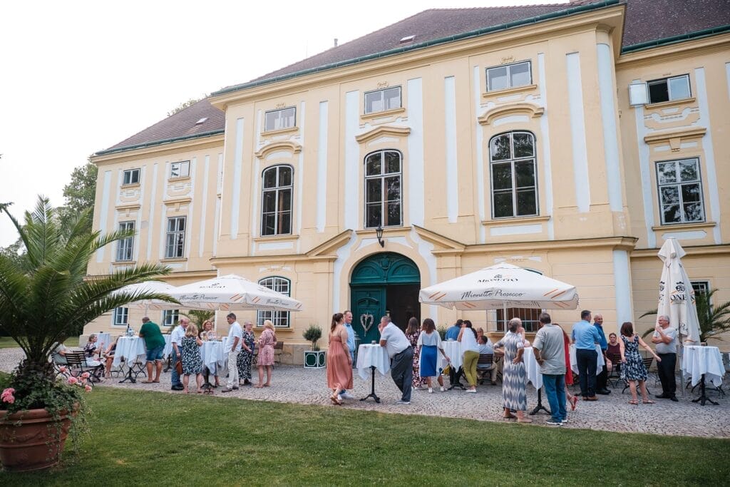 Hochzeitskrone Menschen unterhalten sich bei einer Veranstaltung im Freien vor einem großen, cremefarbenen Gebäude mit grünen Türen und Bogenfenstern – die Essenz solcher Momente einzufangen, ist die Kunst eines Hochzeitsfotografen in Wien und Niederösterreich. So sorgt er dafür, dass jedes Bild seine eigene Geschichte erzählt.