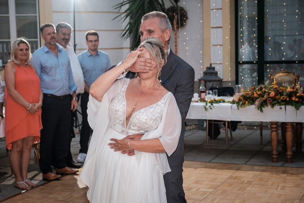 Hochzeitskrone Ein Paar tanzt auf einer Hochzeit, der Mann bedeckt spielerisch die Augen der Frau. Die Zuschauer schauen von der Seite zu und halten echte Momente fest. Ein dekorierter Tisch ziert die Kulisse, perfekt für authentische Hochzeitsfotos, aufgenommen von einem erfahrenen Hochzeitsfotografen in Wien und Niederösterreich.