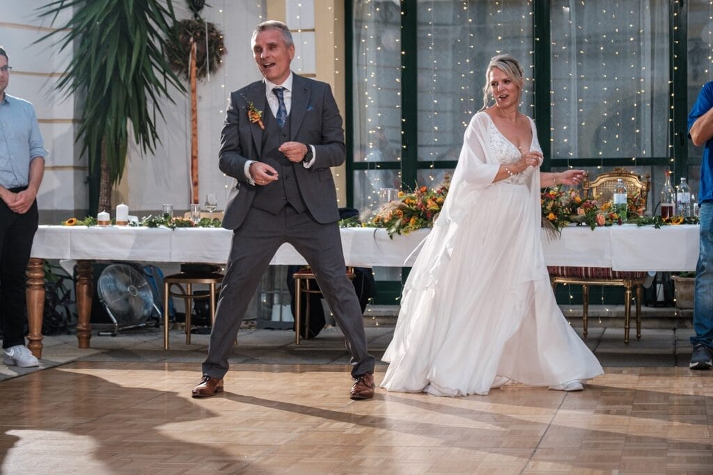 Hochzeitskrone Ein Paar im Hochzeitskleid tanzt begeistert auf einem Holzboden, festgehalten in authentischen Hochzeitsfotos, mit einem dekorierten Tisch und funkelnden Lichterketten im Hintergrund.