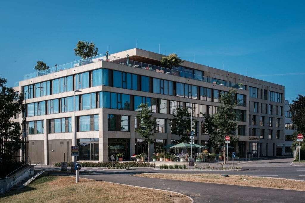 Hochzeitskrone Modernes vierstöckiges Gebäude mit großen Fenstern, umgeben von Bäumen und einem Terrassenbereich mit Sitzgelegenheiten im Freien – eine ideale Kulisse für authentische Hochzeitsfotos. Der klare blaue Himmel im Hintergrund unterstreicht die natürliche Schönheit, perfekt für jeden Hochzeitsfotografen in Wien und Niederösterreich, der malerische Kulissen sucht.