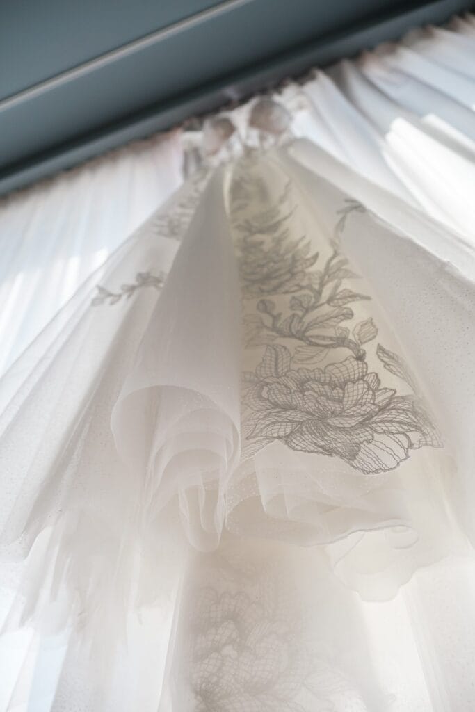 Hochzeitskrone Ein weißes Kleid mit aufwendiger Blumenstickerei hängt vor transparenten Vorhängen und fängt echte Momente voller Eleganz und Schönheit ein.