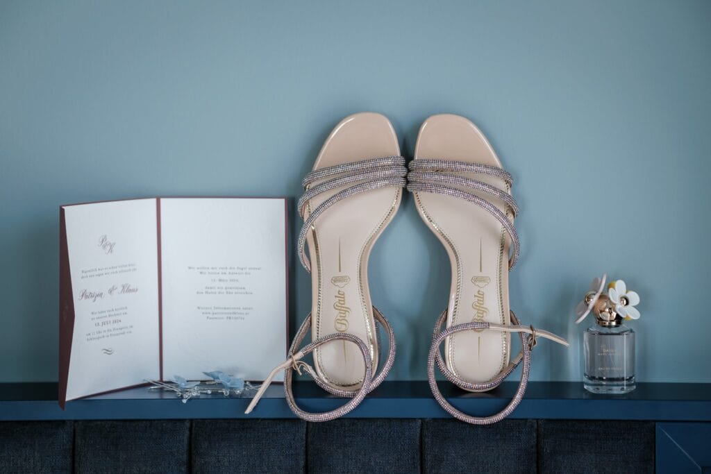 Hochzeitskrone Ein Paar Riemchensandalen, eine Hochzeitseinladung und ein kleines Parfümfläschchen sind ordentlich auf einer dunkelblauen Fläche vor einer hellblauen Wand arrangiert. Sie fangen die Essenz echter Momente ein und bereiten die Bühne für authentische Hochzeitserinnerungen.