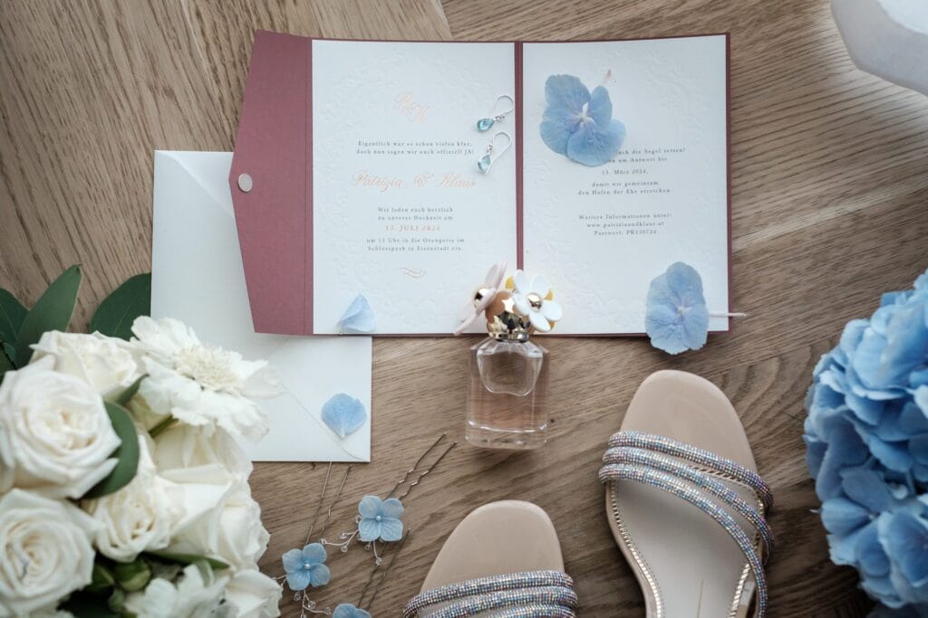 Hochzeitskrone Hochzeitseinladungsset mit floralen Akzenten, einer Parfümflasche, Sandalen und weißen und blauen Blumen, arrangiert auf einer Holzoberfläche – fängt echte Momente für authentische Hochzeitsfotos ein.