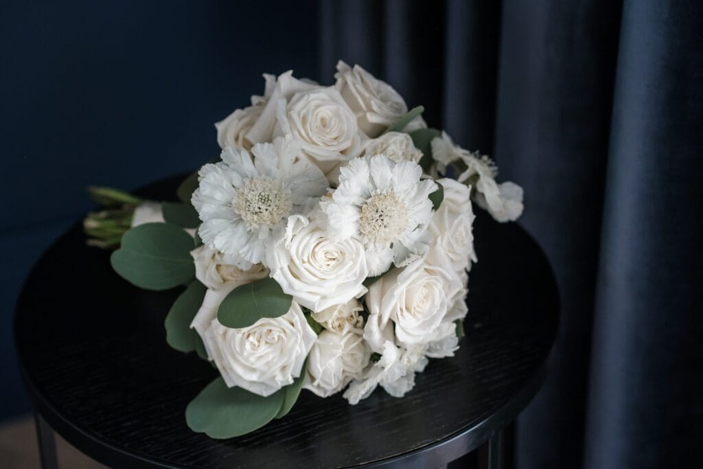 Hochzeitskrone Ein Strauß weißer Rosen und Blumen mit grünen Blättern ziert einen schwarzen runden Tisch vor einem dunklen Vorhanghintergrund – perfekt für die Aufnahme natürlicher Hochzeitsfotos von einem talentierten Hochzeitsfotografen in Wien und Niederösterreich.