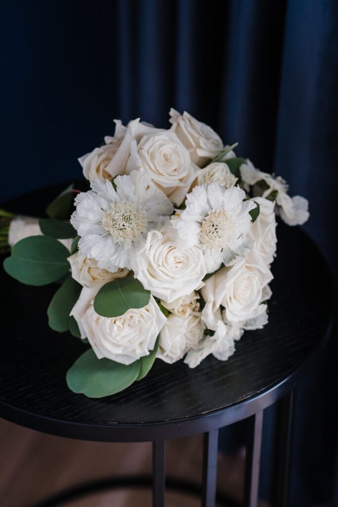 Hochzeitskrone Ein Strauß weißer Rosen und Blumen mit grünen Blättern steht auf einem runden schwarzen Tisch, eingerahmt von einem dunkelblauen Vorhang. Die Szene fängt echte Momente ein und erinnert an natürliche Hochzeitsfotos, die man oft bei Hochzeiten in Wien und Niederösterreich sieht.