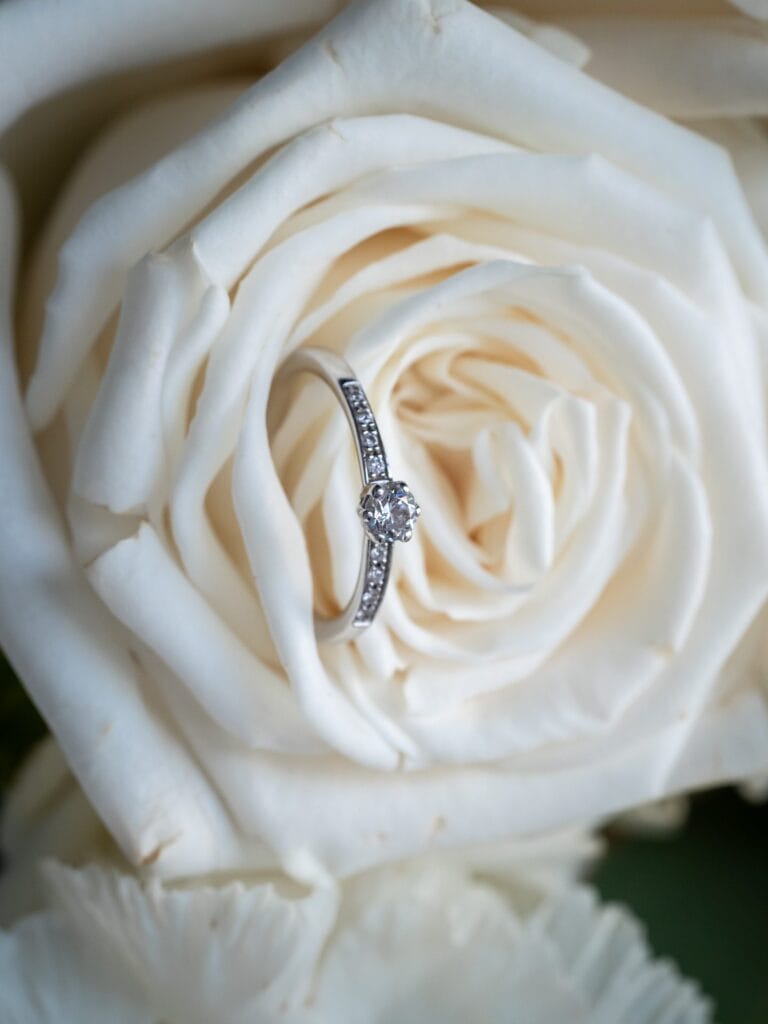 Hochzeitskrone Ein Diamant-Verlobungsring liegt in einer weißen Rose und hält echte Momente fest, die für immer in Erinnerung bleiben.