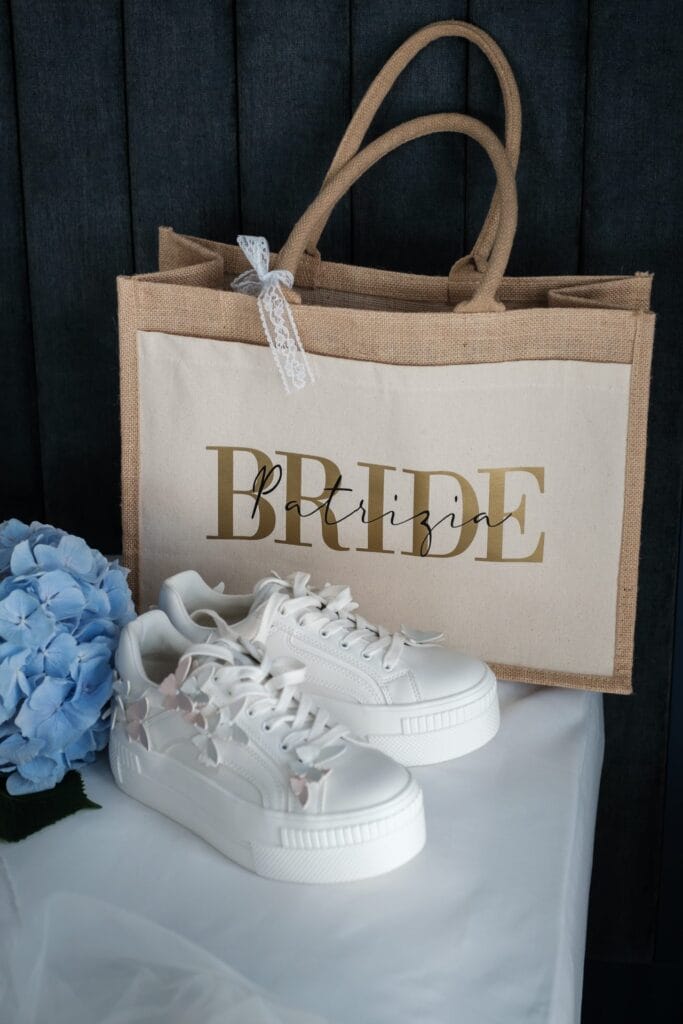 Hochzeitskrone Weiße Sneakers mit floralen Details passen perfekt zu einer „Braut“-Einkaufstasche und einer blauen Hortensienblüte und fangen die Essenz natürlicher Hochzeitsfotos ein.