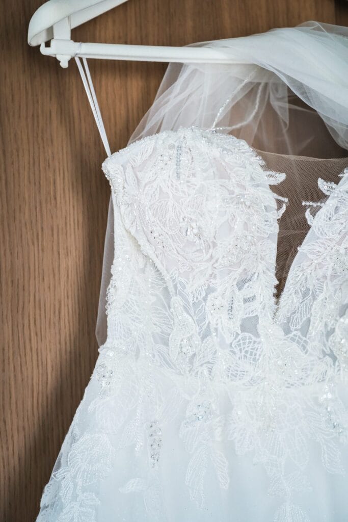 Hochzeitskrone Ein weißes Spitzenbrautkleid mit aufwendiger Blumenstickerei und einem Schleier hängt auf einem hölzernen Kleiderbügel und verkörpert die Eleganz, die in natürlichen Hochzeitsfotos eingefangen wird.