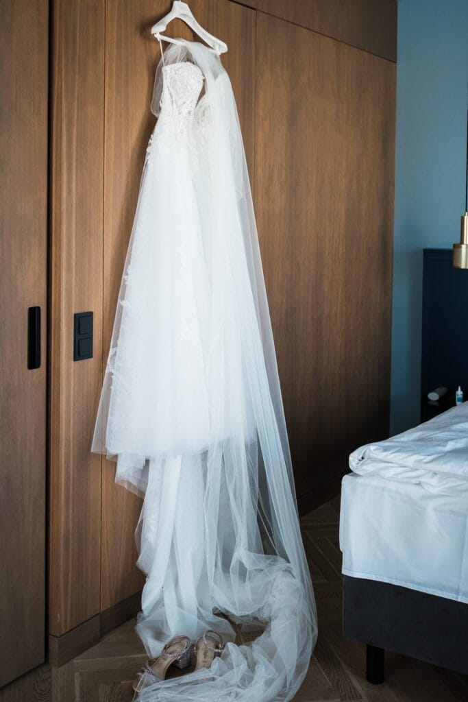 Hochzeitskrone Ein weißes Brautkleid mit langem Schleier hängt anmutig an einer hölzernen Schranktür neben einem Bett, darunter liegt ein Paar Schuhe auf dem Boden und fängt echte Momente ein, gesehen durch die Linse eines Hochzeitsfotografen in Wien und Niederösterreich.