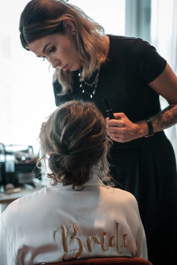 Hochzeitskrone Eine Visagistin schminkt das Gesicht einer Frau, die auf einem Stuhl sitzt. Auf der Rückseite ihres Bademantels steht „Braut“. Dieser Moment fängt die Essenz authentischer Hochzeitsfotos ein und unterstreicht die natürliche Schönheit und Aufregung der Vorbereitungen für den großen Tag.