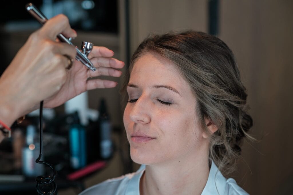 Hochzeitskrone Eine Frau mit geschlossenen Augen erhält Airbrush-Make-up und fängt damit einen dieser echten Momente ein, die authentische Hochzeitsfotos ausmachen. Die Szene entfaltet sich ganz natürlich, wie von einem talentierten Hochzeitsfotografen in Wien und Niederösterreich eingerahmt.