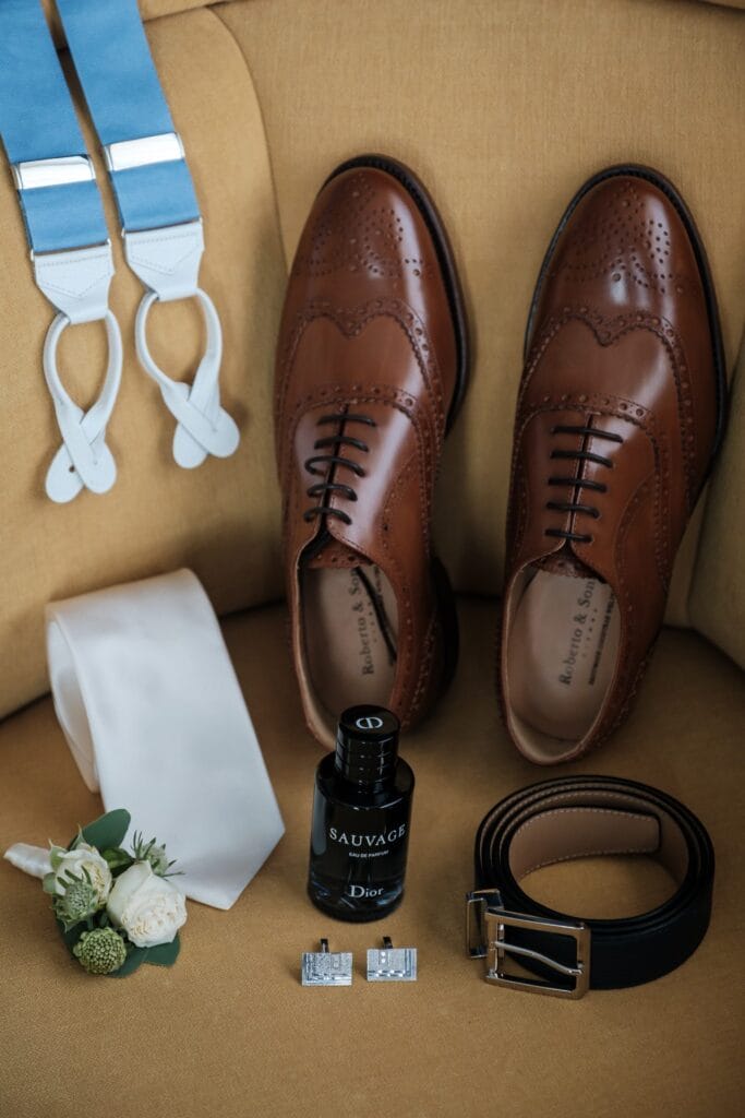 Hochzeitskrone Braune Lederschuhe, ein schwarzer Gürtel, eine weiße Krawatte, eine Flasche Kölnisch Wasser, eine Boutonniere, Hosenträger und Manschettenknöpfe liegen auf einem hellbraunen Stuhl – perfekte Details für authentische Hochzeitsfotos, die die Essenz Ihres besonderen Tages einfangen.