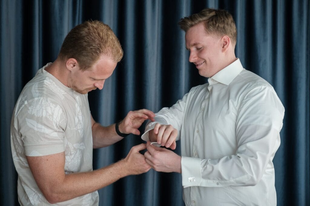 Hochzeitskrone Zwei Männer stehen vor einem dunklen Vorhang, einer hilft dem anderen aufmerksam beim Zurechtknöpfen seiner Hemdmanschetten. Ein Hochzeitsfotograf in Wien und Niederösterreich fängt diesen Moment ein und zeigt die Anmut und Authentizität, die oft in natürlichen Hochzeitsfotos zu finden ist.