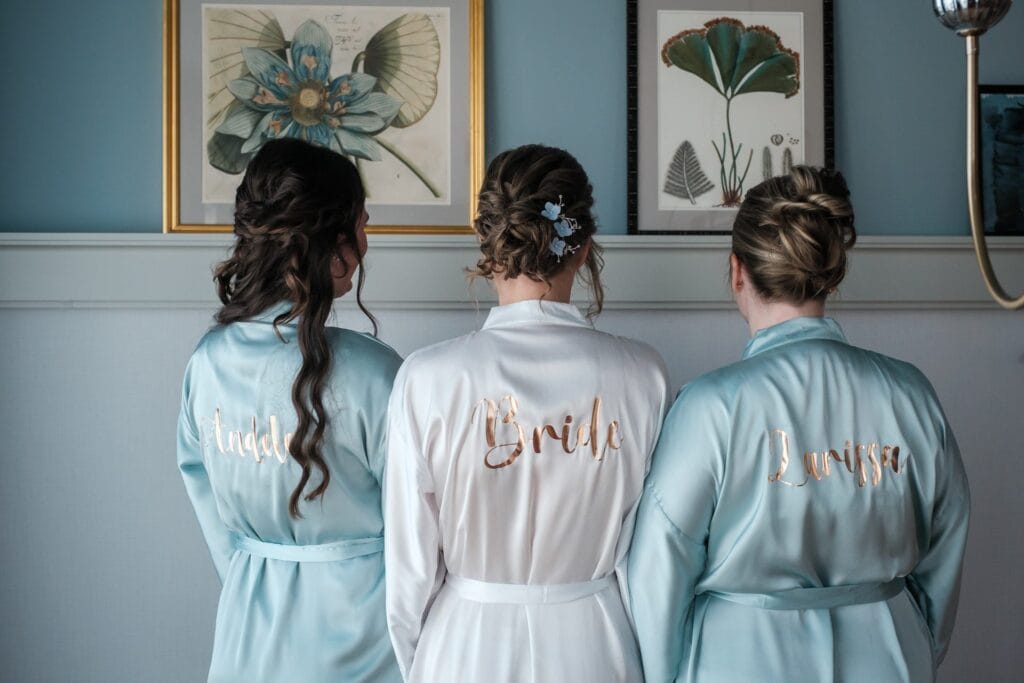 Hochzeitskrone Drei Frauen in Roben, eine mit der Aufschrift „Braut“ in Weiß, stehen abgewandt. Die anderen tragen hellblaue Roben mit „Linda“ und „Louisa“ in Gold. Botanische Drucke hängen an einer blauen Wand – sie fangen die Essenz natürlicher Hochzeitsfotos für einen Hochzeitsfotografen in Wien und Niederösterreich perfekt ein.