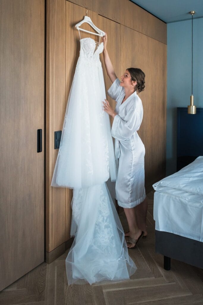 Hochzeitskrone Eine Frau im Morgenmantel steht neben einem Holzschrank und hält ein weißes Brautkleid auf einem Kleiderbügel hoch. Damit fängt sie echte Momente ein, die sich perfekt für natürliche Hochzeitsfotos eignen.