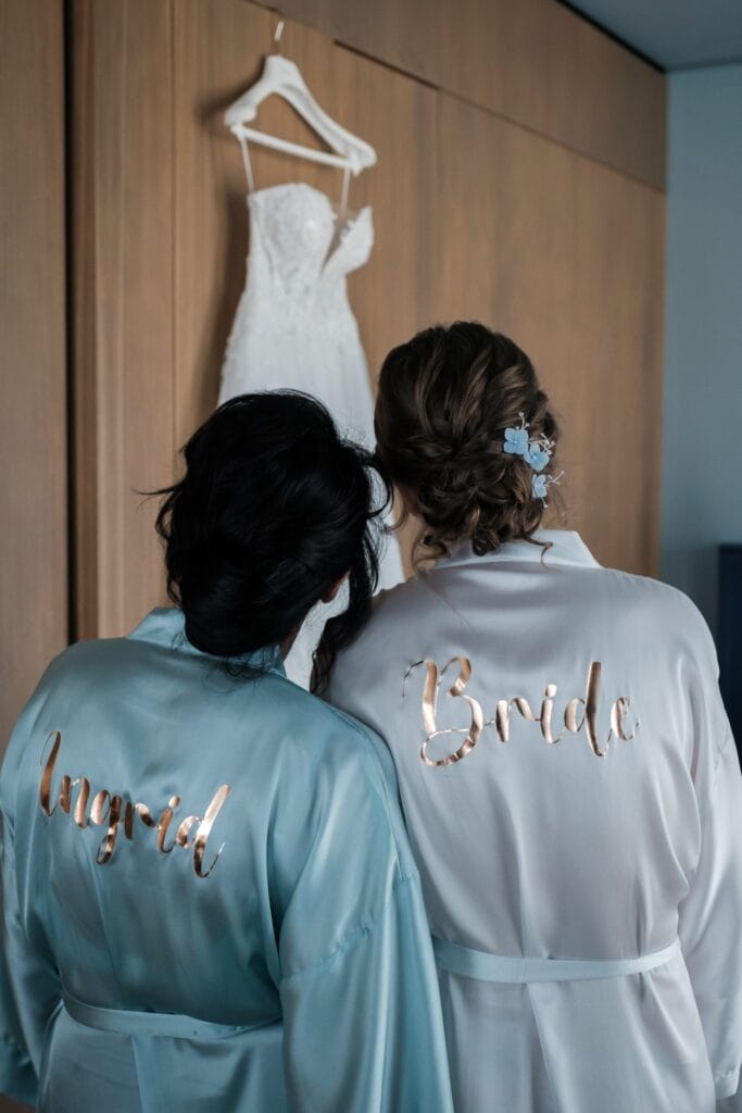 Hochzeitskrone Zwei Frauen in Roben mit „Ingrid“ und „Braut“ auf dem Rücken bewundern ein an einer Holzwand hängendes Brautkleid und fangen echte Momente ein, die für authentische Hochzeitsfotos sorgen.