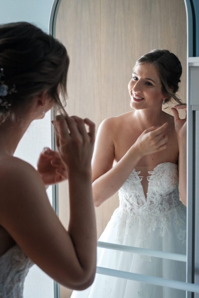 Hochzeitskrone Eine Braut in einem weißen Spitzenbrautkleid richtet ihr Haar, während sie in den Spiegel schaut, und fängt so echte Momente ein, die perfekt für authentische Hochzeitsfotos sind.