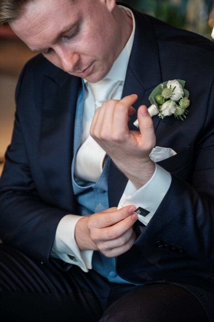 Hochzeitskrone Ein Mann im Anzug rückt seine Manschettenknöpfe zurecht, eine zarte Boutonnière ziert sein Jackett. Aufgenommen von einem Hochzeitsfotografen in Wien und Niederösterreich, verkörpert dieses Bild die Eleganz und Essenz authentischer Hochzeitsmomente.