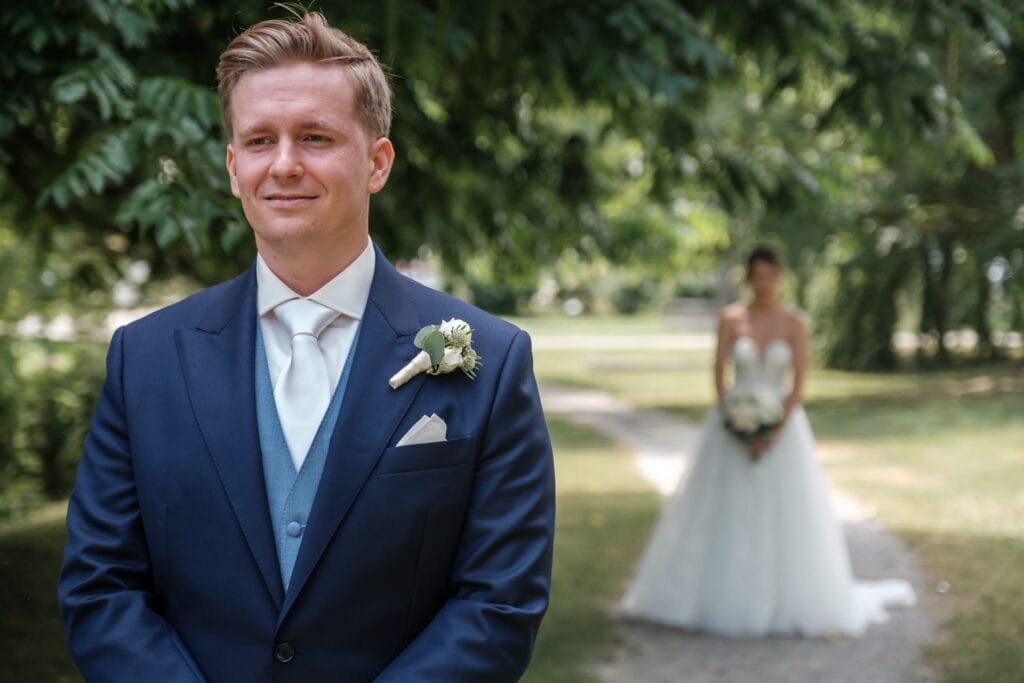 Hochzeitskrone Ein Bräutigam im blauen Anzug steht im Vordergrund, während die Braut in einem weißen Kleid mit Blumenstrauß sanft verschwommen auf einem von Bäumen gesäumten Weg steht. Diese Szene fängt natürliche Hochzeitsfotos von einem Hochzeitsfotografen in Wien und Niederösterreich ein.