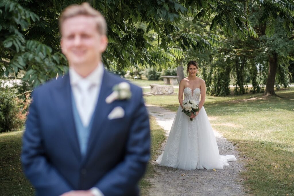 Hochzeitskrone Eine Braut im weißen Kleid hält einen Blumenstrauß und fängt echte Momente ein, während der Bräutigam im blauen Anzug im Vordergrund unter üppigen grünen Bäumen verschwommen ist.