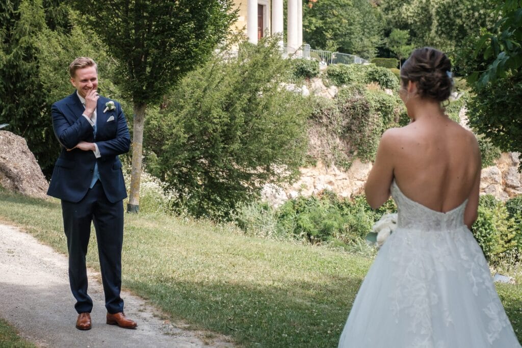 Hochzeitskrone Ein Bräutigam im blauen Anzug, der „echte Momente“ verkörpert, lächelt mit der Hand am Kinn, während er im Freien in der Nähe eines Gebäudes und von Bäumen eine Braut im weißen Kleid ansieht.