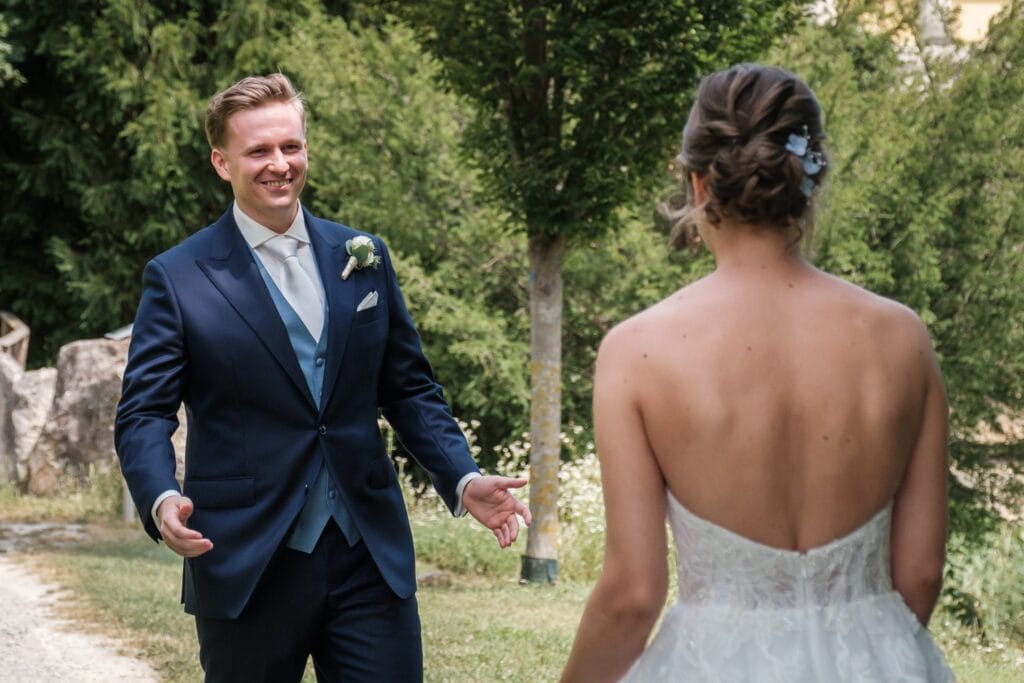 Hochzeitskrone Ein Mann im Anzug lächelt und nähert sich einer Frau in einem schulterfreien weißen Kleid im Freien auf einem von Grün umgebenen Weg und fängt echte Momente ein. Perfekt für natürliche Hochzeitsfotos, diese bezaubernde Szene ist der Traum eines jeden Hochzeitsfotografen in Wien und Niederösterreich.