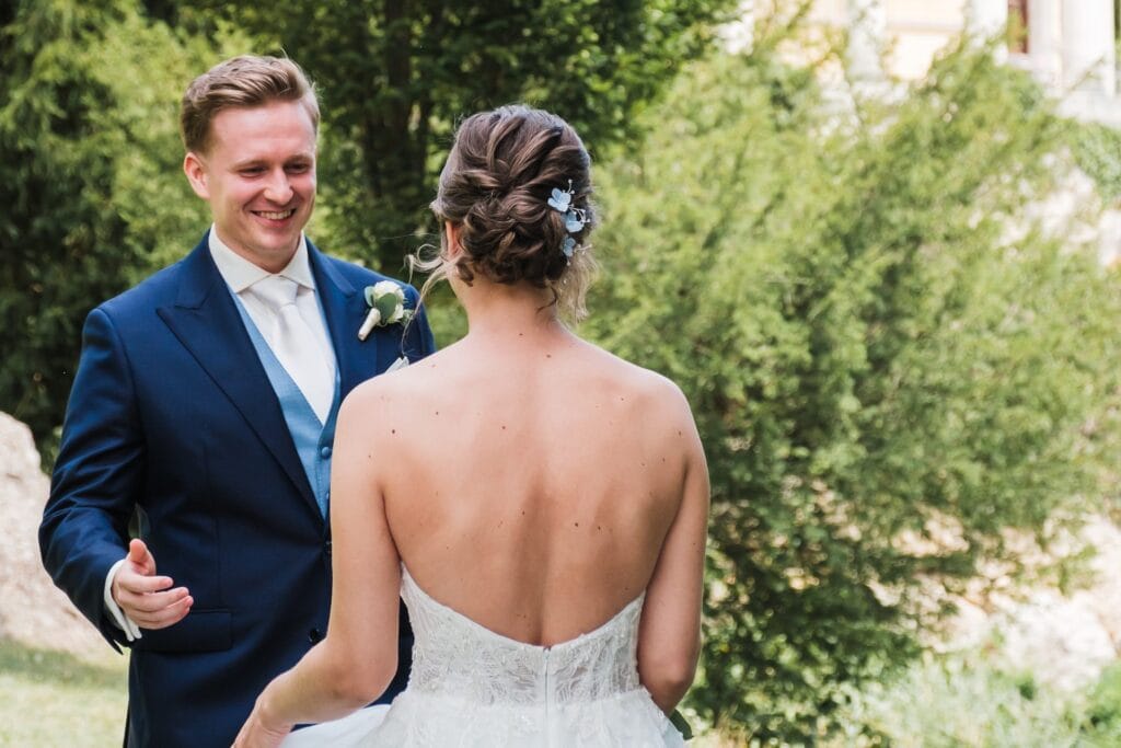 Hochzeitskrone Ein Mann im blauen Anzug lächelt eine Frau in einem weißen trägerlosen Kleid an, umgeben von üppigem Grün. Diese bezaubernde Szene fängt echte Momente ein und eignet sich perfekt für eine authentische Hochzeitsfotos-Sammlung von einem Hochzeitsfotografen in Wien und Niederösterreich.