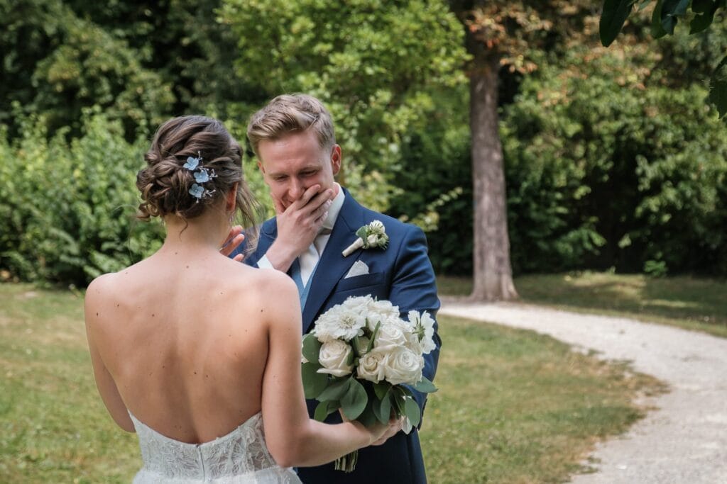 Hochzeitskrone Der Bräutigam reagiert emotional, als er die Braut in ihrem weißen Kleid mit einem Blumenstrauß in der Hand vor einer üppigen Kulisse im Freien sieht. Eingefangen von einem Hochzeitsfotografen in Wien und Niederösterreich ist dies einer jener echten Momente, die natürliche Hochzeitsfotos wirklich unvergesslich machen.