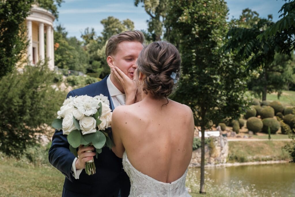 Hochzeitskrone Braut und Bräutigam umarmen sich im Freien und halten echte Momente fest. Die Braut in einem schulterfreien Kleid mit weißen Rosen in den Händen wiegt sanft das Gesicht des Bräutigams. Er steht elegant in einem Anzug da. Grünflächen und ein Gebäude rahmen diese Szene authentischer Hochzeitsfotos eines Hochzeitsfotografen in Wien und Niederösterreich ein.