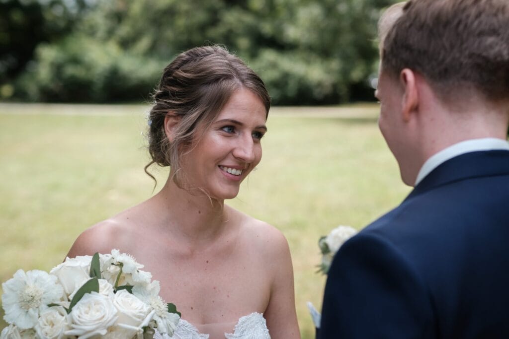 Hochzeitskrone Braut und Bräutigam stehen im Freien und lächeln sich in einer bezaubernden Ausstellung natürlicher Hochzeitsfotos an. Die Braut hält einen Strauß weißer Blumen in den Händen, umgeben von üppigem Grün, das die perfekte Kulisse für ihre Liebesgeschichte bildet.