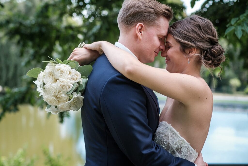 Hochzeitskrone Braut und Bräutigam umarmen sich und berühren sich mit den Stirnen, um echte Momente festzuhalten. Die Braut hält sanft einen Strauß weißer Rosen, während sie draußen neben einem ruhigen Gewässer stehen und authentische Hochzeitsfotos voller Liebe und natürlicher Schönheit entstehen lassen.