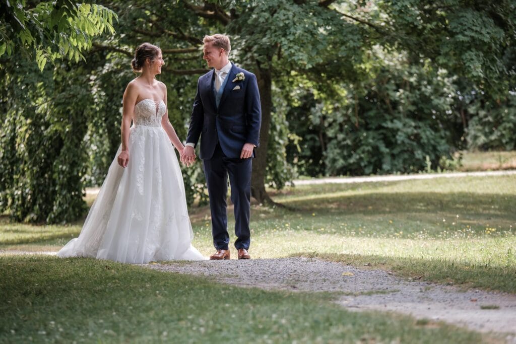 Hochzeitskrone Braut und Bräutigam stehen Händchen haltend auf einem von Bäumen umgebenen Grasweg und machen authentische Hochzeitsfotos von echten Momenten.