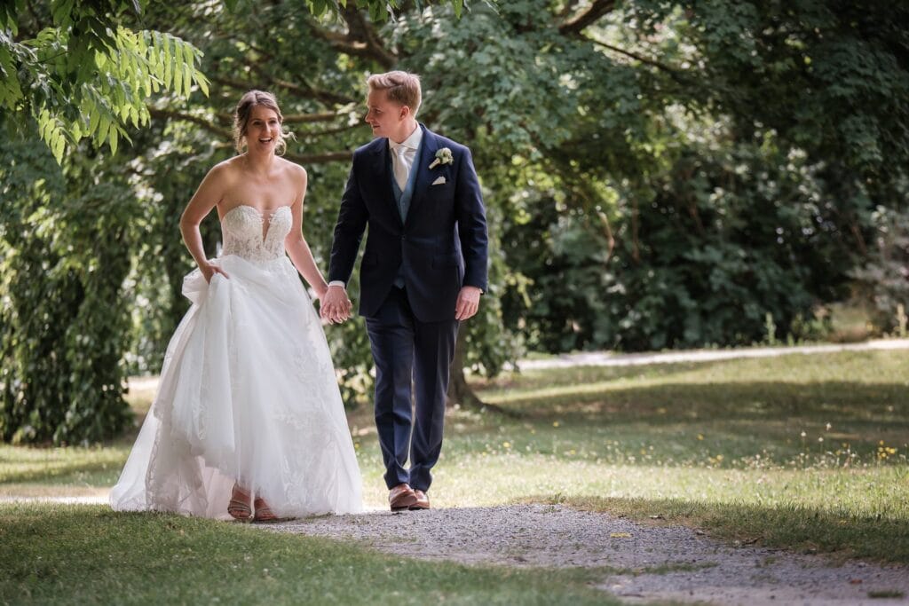 Hochzeitskrone Eine Braut und ein Bräutigam gehen Hand in Hand auf einem von Grün umgebenen Kiesweg und fangen die Essenz authentischer Hochzeitsfotos ein.