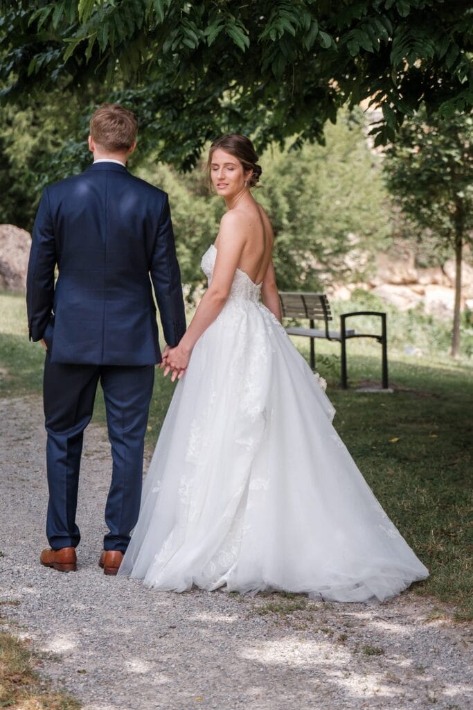 Hochzeitskrone Ein Paar geht Händchen haltend auf einem Kiesweg. Die Frau trägt ein schulterfreies weißes Kleid und der Mann einen dunklen Anzug. Sie sind draußen, mit Bäumen und einer Bank in der Nähe, und fangen echte Momente ein, die nur ein Hochzeitsfotograf in Wien und Niederösterreich wirklich einfangen kann.