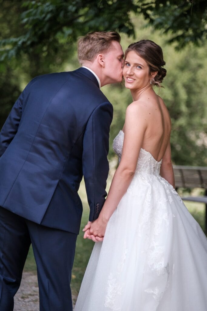 Hochzeitskrone Ein Paar in formeller Kleidung, der Mann küsst die Wange der Frau, hält Händchen und posiert im Freien in einer grünen Umgebung. Der Moment fängt die Essenz natürlicher Hochzeitsfotos ein, perfekt für alle, die einen authentischen Hochzeitsfotografen in Wien und Niederösterreich suchen.