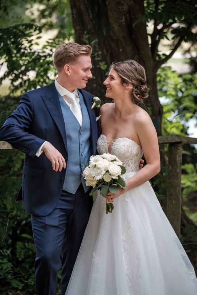 Hochzeitskrone Braut und Bräutigam stehen dicht beieinander im Freien, aufgenommen von einem Hochzeitsfotografen in Wien und Niederösterreich. Die Braut hält einen Strauß weißer Blumen und der Bräutigam trägt einen dunklen Anzug mit einer hellblauen Weste. Beide lächeln sich an und spiegeln echte Momente wider.